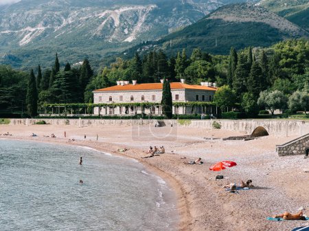 Téléchargez les photos : Les touristes prennent un bain de soleil sur la plage près de la Villa Milocer au pied des montagnes. Monténégro. Photo de haute qualité - en image libre de droit