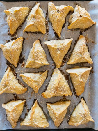 Triangular puffs with filling sprinkled with sesame seeds lie on a baking sheet. High quality photo