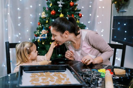Foto de La pequeña hija trata a su madre con una galleta mientras está sentada en una mesa con galletas crudas en una bandeja. Foto de alta calidad - Imagen libre de derechos