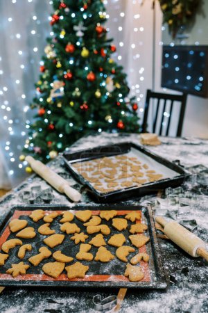 Foto de Bandejas para hornear con galletas de jengibre crudas y horneadas sobre una mesa enharinada cerca del árbol de Navidad. Foto de alta calidad - Imagen libre de derechos