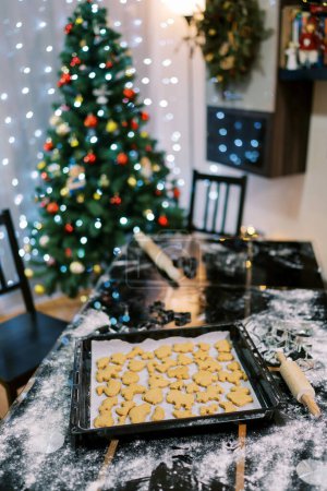 Foto de Las galletas de Ruddy mienten en el papel de hornear en una bandeja en una tabla enharinada al lado del árbol de Navidad. Foto de alta calidad - Imagen libre de derechos