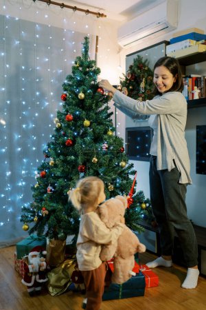 Téléchargez les photos : Une petite fille avec un ours en peluche donne le sommet du sapin de Noël à sa mère accrochant des boules. Photo de haute qualité - en image libre de droit