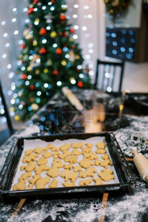 Foto de Las galletas cocidas se encuentran sobre el papel de hornear en la bandeja sobre la mesa cerca del árbol decorado de Navidad. Foto de alta calidad - Imagen libre de derechos