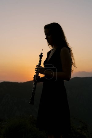 Fille avec une clarinette dans les mains se tient dans les montagnes au coucher du soleil. Photo de haute qualité