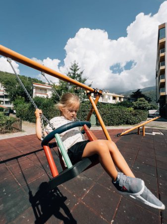 Foto de La niña se balancea en un columpio de cadena en el patio de recreo cerca de las casas. Foto de alta calidad - Imagen libre de derechos