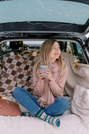Téléchargez les photos : Jeune femme souriante avec tasse de café assise dans le coffre de la voiture et regardant ailleurs. Photo de haute qualité - en image libre de droit