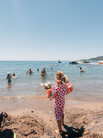 Photo for Little girl in a swimsuit with inflatable armlets stands by the sea and looks at people bathing. High quality photo - Royalty Free Image
