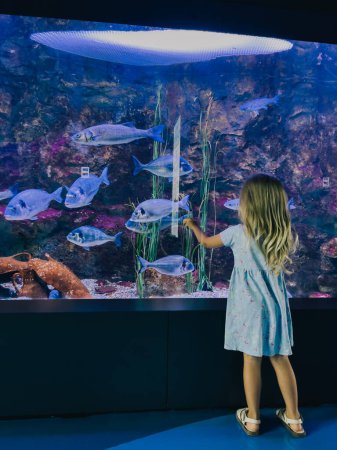 Foto de La niña toca el vaso de un acuario grande con peces flotantes con sus manos. Foto de alta calidad - Imagen libre de derechos