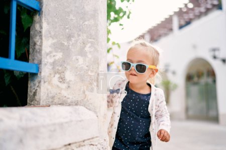 Téléchargez les photos : La petite fille aux lunettes de soleil jette un coup d'oeil par derrière une clôture dans la cour. Photo de haute qualité - en image libre de droit