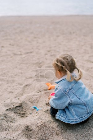 Téléchargez les photos : Petite fille assise sur une plage de sable avec des moules colorés et des pelles. Vue de derrière. Photo de haute qualité - en image libre de droit