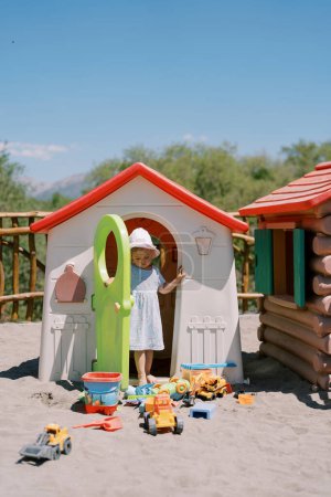 Téléchargez les photos : Une petite fille sort d'une maison de jouets et regarde les jouets sur le sable. Photo de haute qualité - en image libre de droit