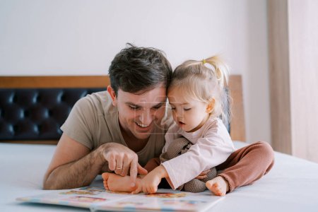 Téléchargez les photos : Papa et petite fille lisent un livre de contes de fées sur le lit, passant leurs doigts à travers les pages. Photo de haute qualité - en image libre de droit
