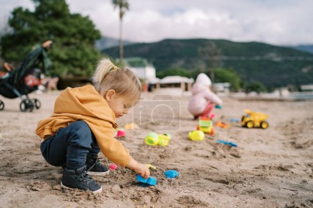 Téléchargez les photos : Petite fille squats sur la plage et joue avec des moules de sable en plastique. Photo de haute qualité - en image libre de droit