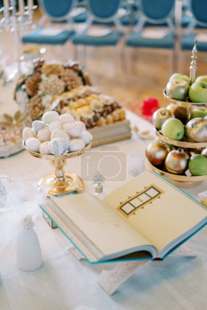 Téléchargez les photos : Ouvrez un livre sacré sur un stand sur une table à côté de fruits dans des vases, des pâtisseries, des cônes de sucre et des noix. Tradition de Sofreh Aghd. Photo de haute qualité - en image libre de droit
