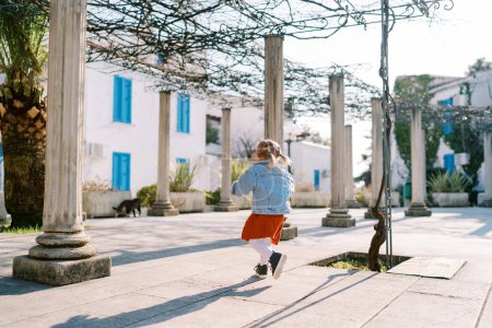 Little girl walks between pergola columns in a garden near a three-story building. High quality photo