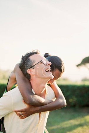 Laughing man carries woman on his back across green lawn. High quality photo