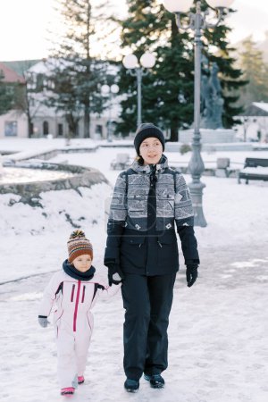 Mom and little girl walk holding hands through a snowy park. High quality photo