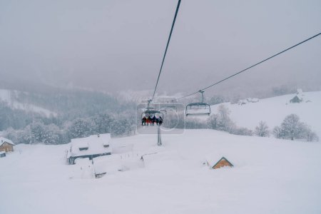 Skiers ride a chairlift up the mountain to snow-covered cottages among a coniferous forest. High quality photo