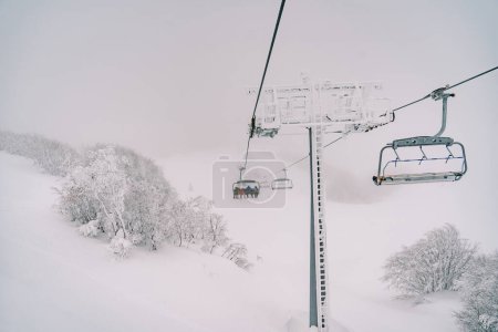Chairlift with skiers rides over snow-covered trees to misty mountain. High quality photo
