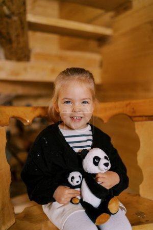 Téléchargez les photos : Petite fille souriante assise avec des pandas de jouets mous sur une chaise en bois. Photo de haute qualité - en image libre de droit