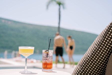 Two cocktails in glasses standing on a table near a sun lounger on the beach against the backdrop of walking man and woman. High quality photo