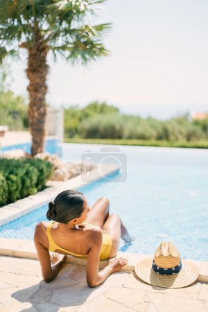 Young woman sunbathes while sitting on the edge of the pool, leaning on her elbows. Back view. High quality photo