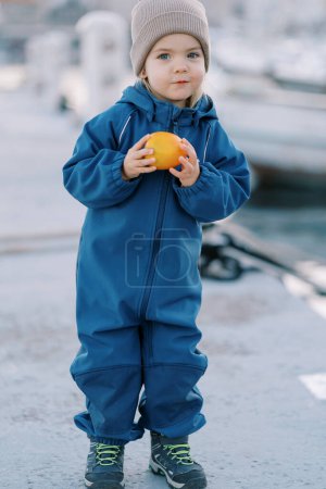 Little smiling girl in overalls with ripe persimmon stands on the pier. High quality photo