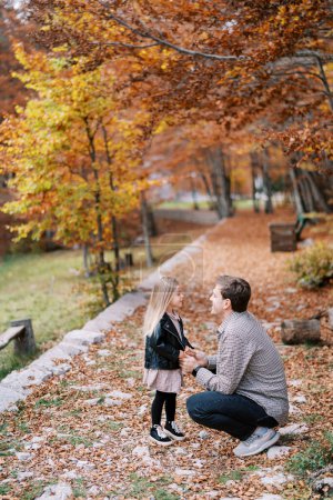 Dad squatted in front of the little girl holding her hands on a rocky path in the autumn forest. High quality photo