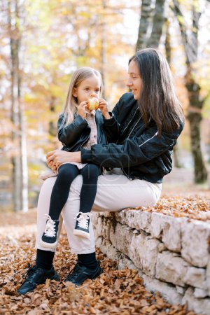Téléchargez les photos : Une petite fille ronge une pomme alors qu'elle est assise dans les bras de sa mère sur le trottoir d'un parc d'automne. Photo de haute qualité - en image libre de droit