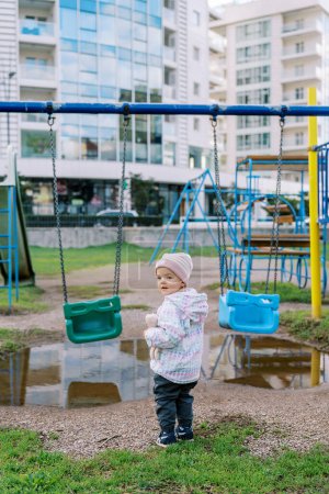 Little girl stands near the swing in front of a puddle, turning around. Back view. High quality photo