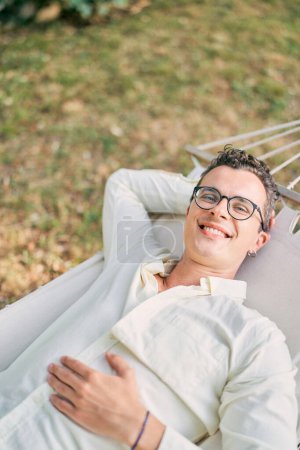 Junger lächelnder Mann liegt mit der Hand unter dem Kopf in einer Hängematte im Garten. Hochwertiges Foto