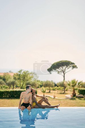 Frau lehnt am Rande eines Swimmingpools in einem grünen Garten an Mann. Hochwertiges Foto