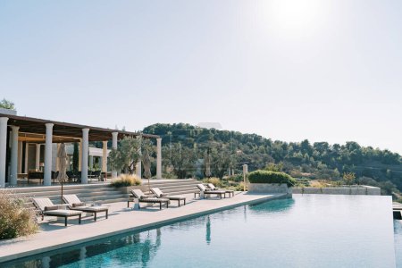 Sonnenliegen mit zusammengefalteten Sonnenschirmen säumen die Säulenterrasse neben dem langen Swimmingpool. Hotel Amanzoe. Peloponnes, Griechenland. Hochwertiges Foto