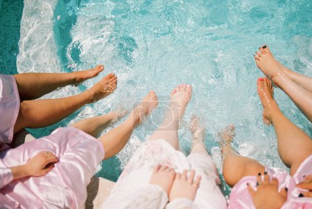 Bride and her bridesmaids sit on the edge of the pool with their feet in the turquoise water. Cropped. Faceless. High quality photo