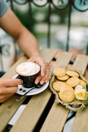 Mann hält eine Tasse Cappuccino mit den Händen, während er an einem Tisch auf der Terrasse neben einem Tablett mit Keksen sitzt. Beschnitten. Gesichtslos. Hochwertiges Foto