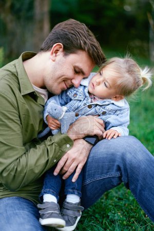 Papa umarmt ein kleines Mädchen, das auf den Knien im Park sitzt. Hochwertiges Foto