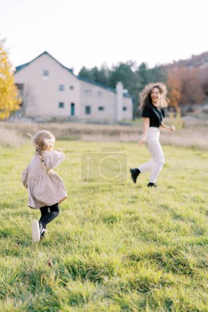 Little girl runs after her laughing mother across the green lawn. Back view. High quality photo