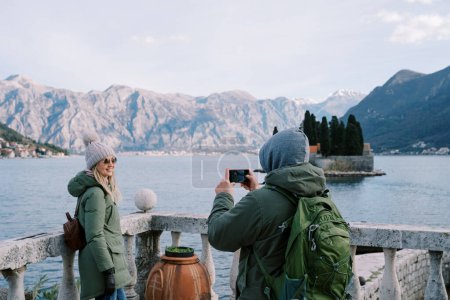 Guy macht ein Foto von Mädchen mit einem Smartphone in der Nähe der Brüstung am Ufer der Bucht von Kotor mit Blick auf die Insel St. George. Montenegro. Rückansicht. Hochwertiges Foto