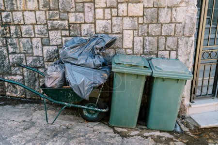 Cart with bags of garbage stands near plastic garbage containers near a stone fence. High quality photo