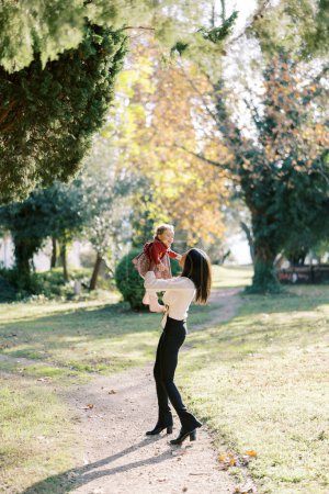 Smiling mother circling little girl in her arms while standing on path in park. High quality photo
