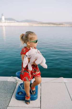 Little girl with a pink soft toy rabbit sits on a bollard on the pier and looks away. High quality photo