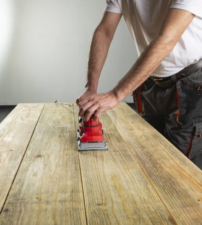 Foto de Máquina de lijar en mano masculina. Procesamiento de la pieza de trabajo en mesa de madera marrón claro. Taller de carpintería - Imagen libre de derechos
