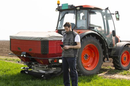 Junge Landarbeiterin neben Traktor mit digitalem Tablet Traktor verteilt Kunstdünger im Weizenfeld. Verkehr, Landwirtschaft.