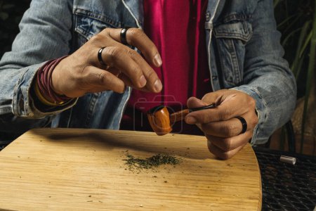 Mann bereitet seine Pfeife mit Marihuana auf einem Holzbrett zu, mit Resten von gemahlenem Marihuana herum.