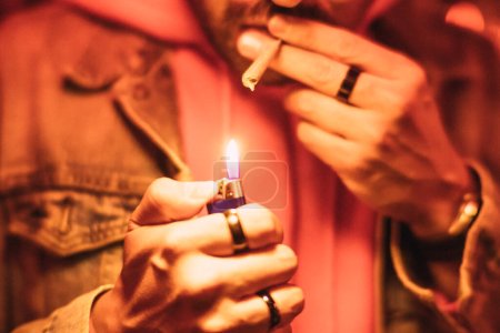 Téléchargez les photos : Détail des mains d'un homme allumant une cigarette de marijuana sous une lumière rouge. - en image libre de droit