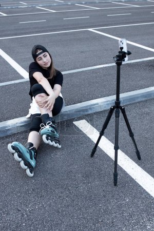 Photo for Girl sitting in an empty parking lot, making selfie. Teenager, rollerblading on her feet, films herself on the front camera of phone on a tripod. Concept for video blog, hobby, content, vlog, blogger. - Royalty Free Image