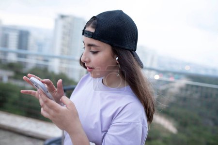 Photo for Stylish caucasian teenager, long hair, black cap, visor back. Young girl is holding a smartphone, typing sms, watching content. Lifestyle, social networks, kids online, communication concept. Outside - Royalty Free Image