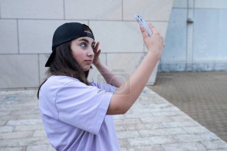 Photo for Teen girl 15 years old with a phone, filming herself on the front camera, makes a selfie. Stylish teenager in a cap. Lifestyle concept, gadget generation, social media life. Copy space. Day light. - Royalty Free Image