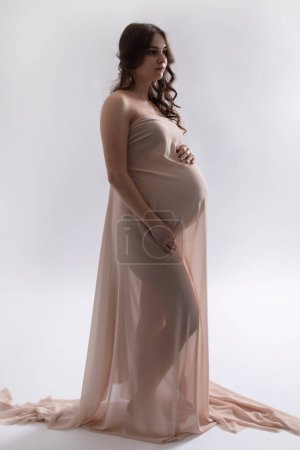 Photo for Beautiful pregnant girl is wrapped in light cloth standing on white studio background. Concept of waiting for baby, 9 months, motherhood, pregnancy. Caucasian woman with long brown hair. - Royalty Free Image