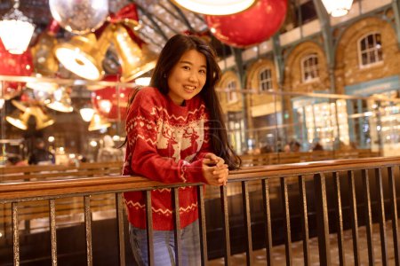 Photo for Cheerful girl with long hair in a Christmas sweater standing in a shopping center. Chinese, young woman 25 years old. Evening. New Year winter fair, holiday concept. - Royalty Free Image